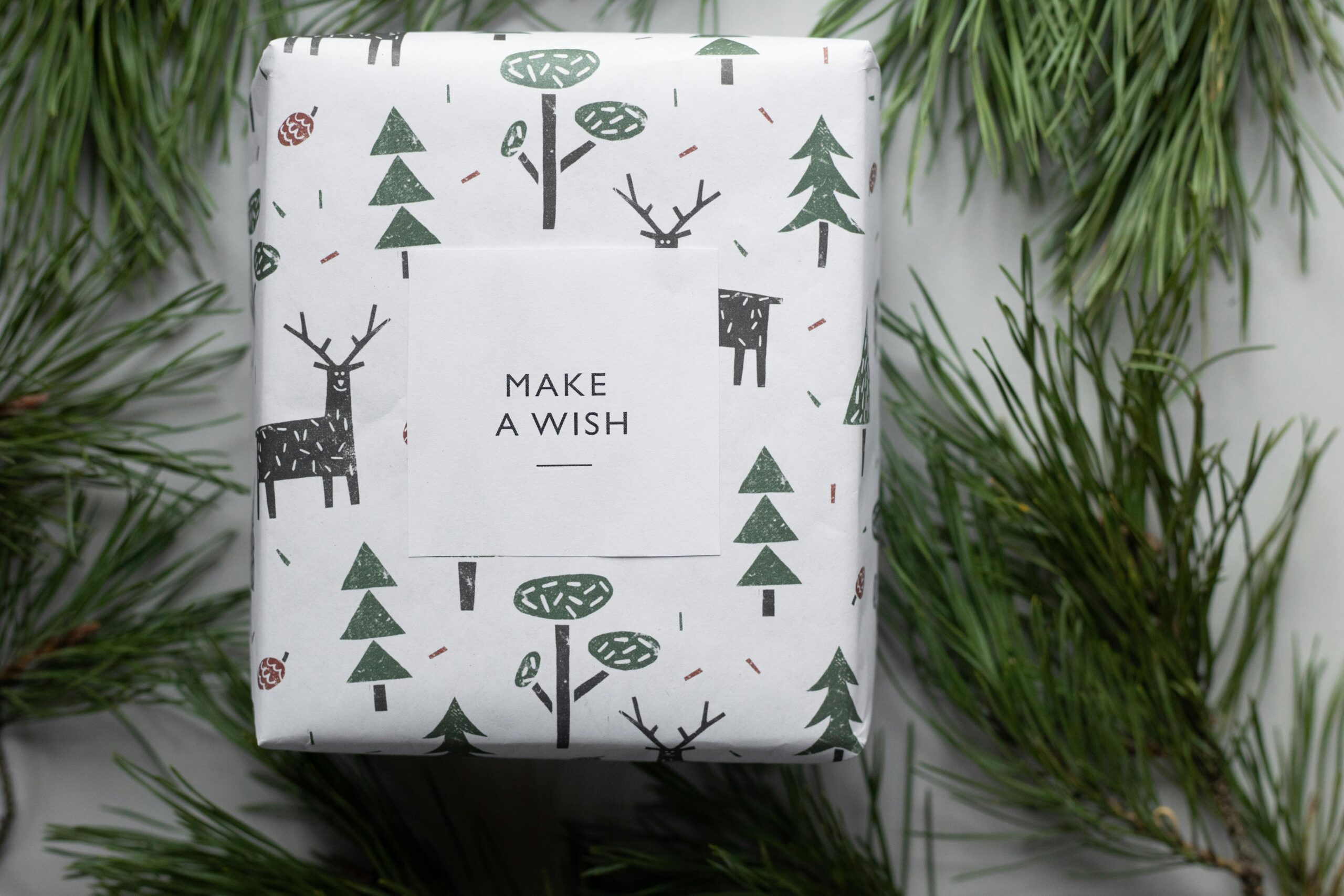 A holiday gift wrapped with tree and deer pattern, surrounded by pine branches, inspiring winter cheer.